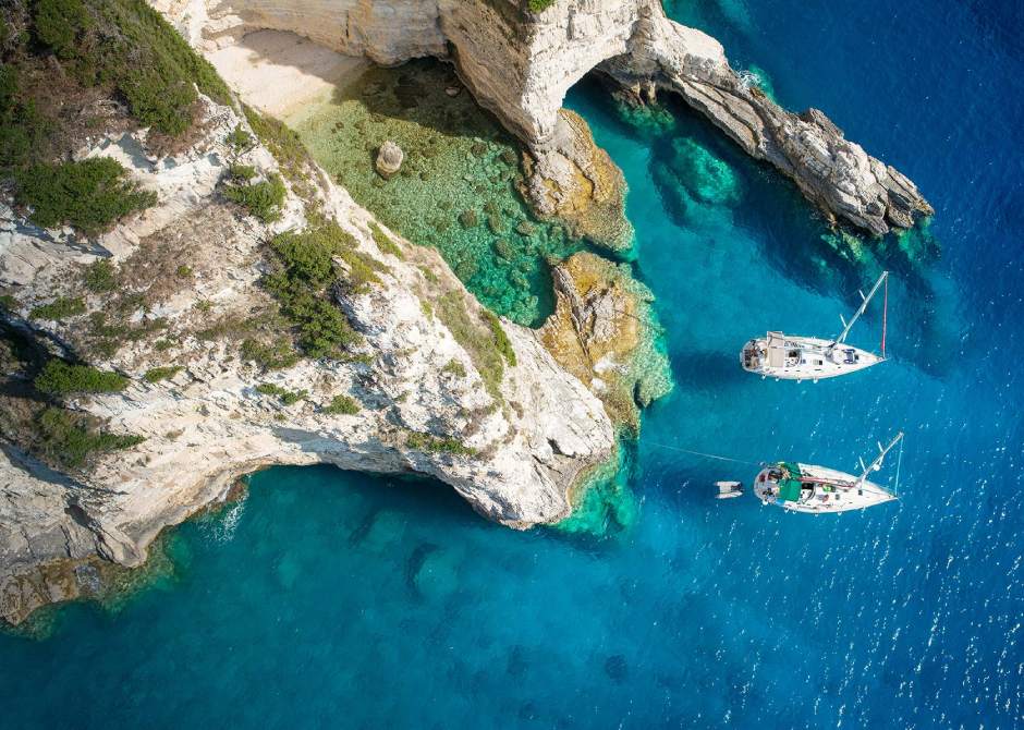 Clear blue waters of Paxi Island, two small boats of Variety Cruises anchored next to a rocky mountain