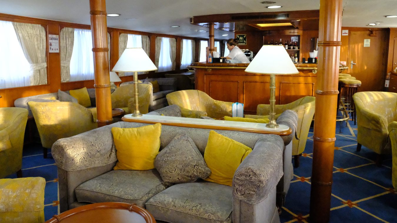 The indoor lounge area of a small cruise ship with comfortable seating, large windows, and elegant decor.